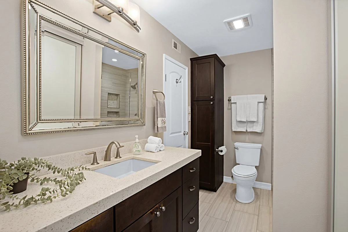 Bathroom with beige walls, a vanity with a white speckled countertop, dark brown cabinets, a large mirror, a small sink, a towel hanger, a dark storage cabinet, and a toilet with organized towels on a wall