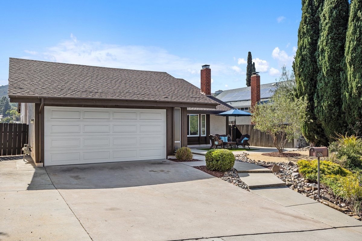 A modern suburban house with a garage, a small front yard with landscaping, a patio area with outdoor furniture and an umbrella, and a mailbox near the sidewalk.