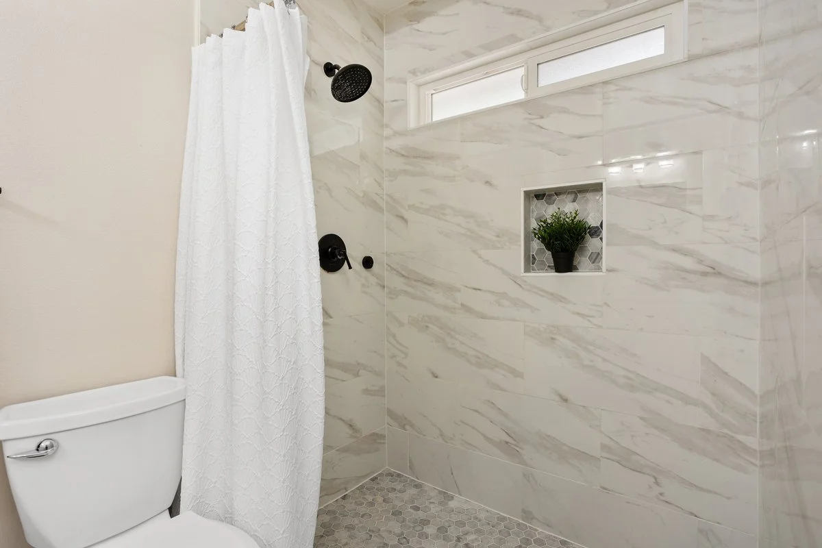 Bathroom shower with black fixtures, a white shower curtain, a small built-in niche with a potted plant, and a frosted window.