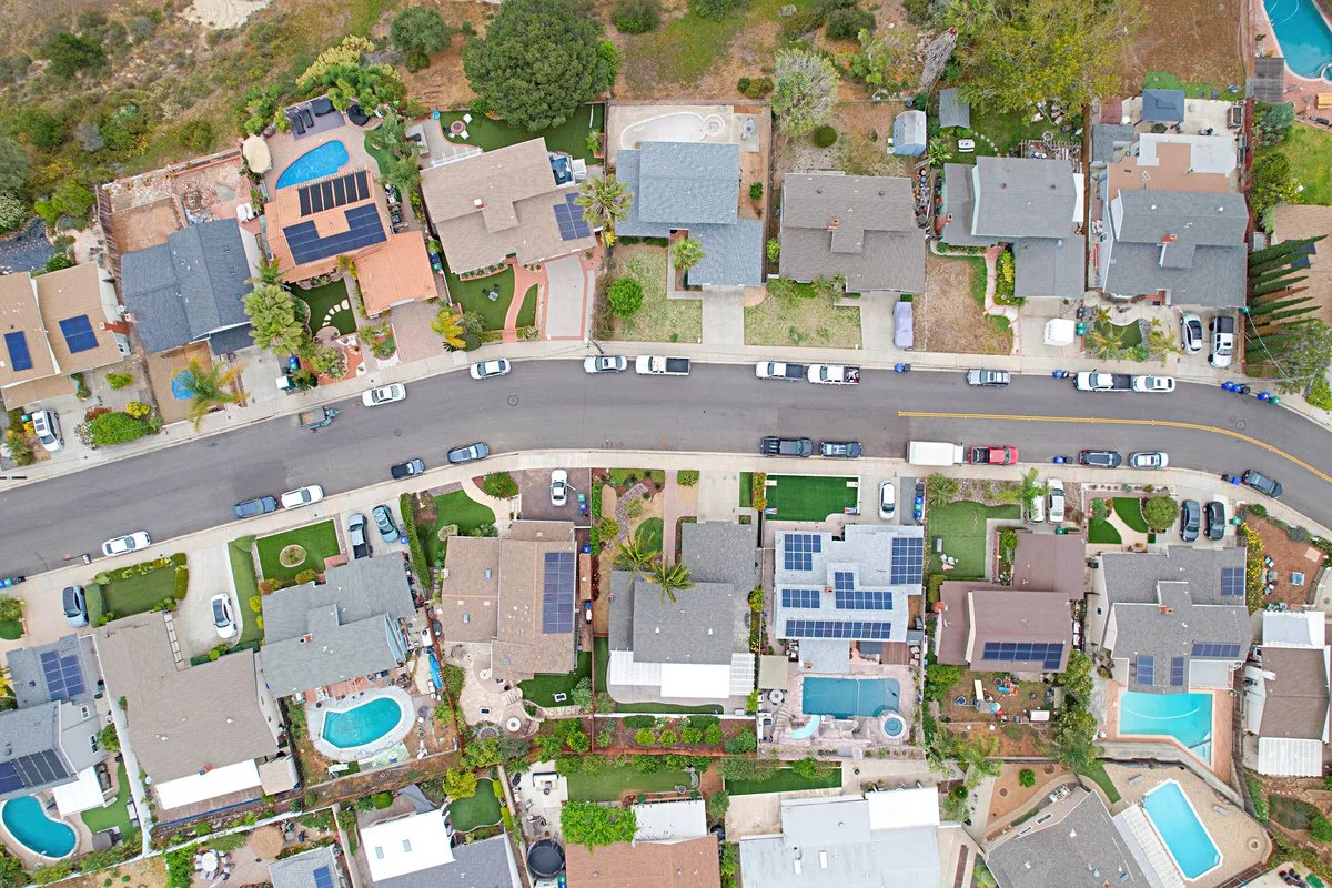 An aerial view of a suburban neighborhood with residential houses that have solar panels on their roofs. Some houses have pools in their backyards, and the street is lined with parked cars.