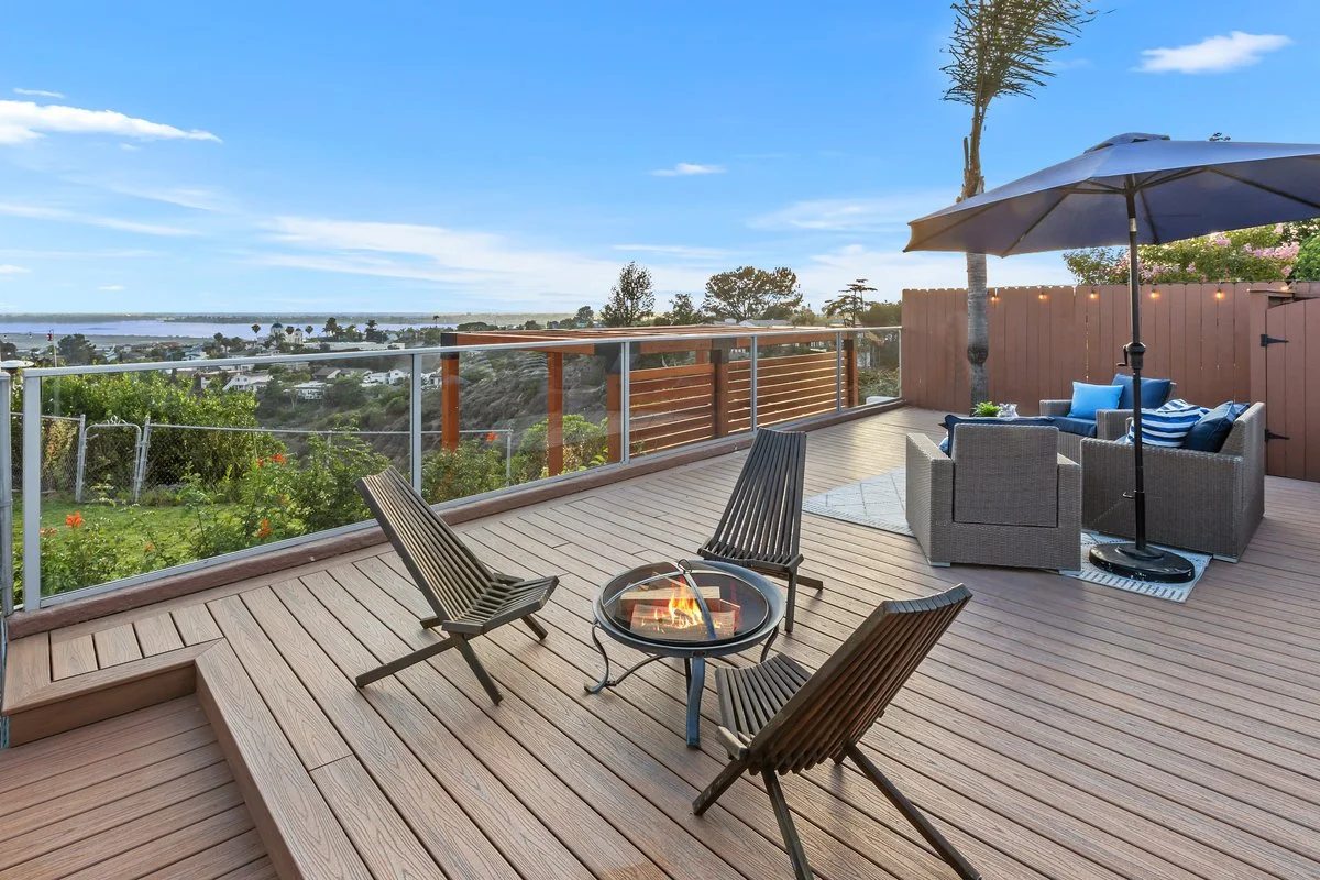 A spacious outdoor wooden deck with seating, including a modern living area with cushioned chairs and an umbrella, overlooking a scenic landscape with trees, water, and a blue sky.