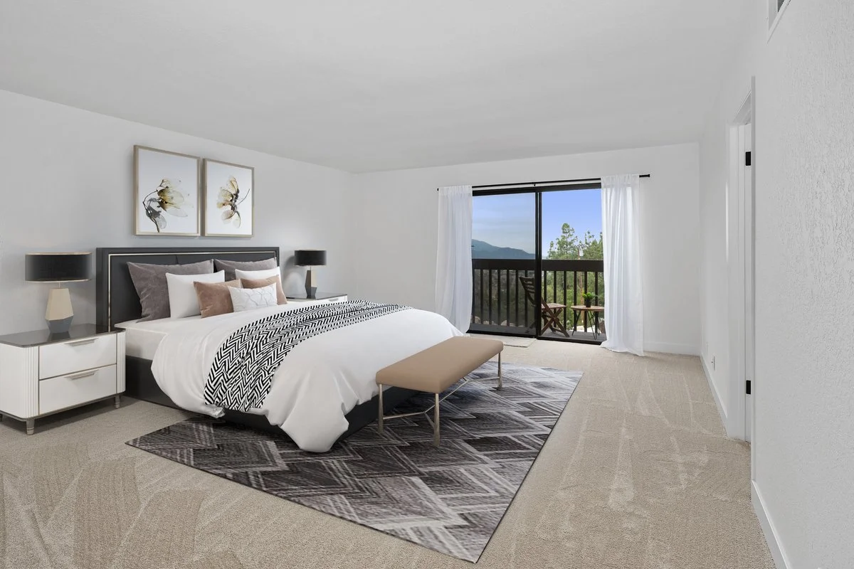 A bedroom with a bed, two nightstands with lamps, and art above the bed. A sliding glass door leads to a balcony with outdoor chairs, overlooking trees and mountains.