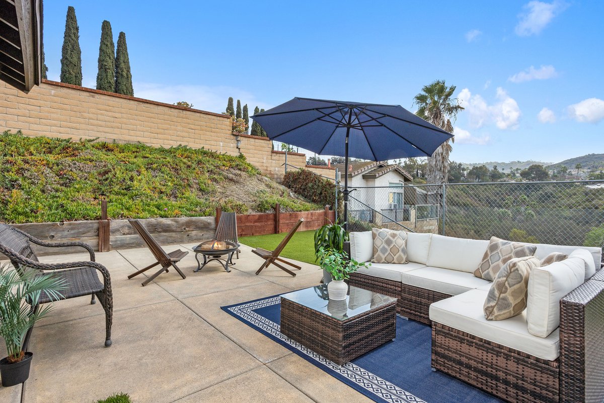 Outdoor patio with beige sectional sofa, blue umbrella, small fire pit, wicker chairs, potted plant, and scenic view of trees and hills.