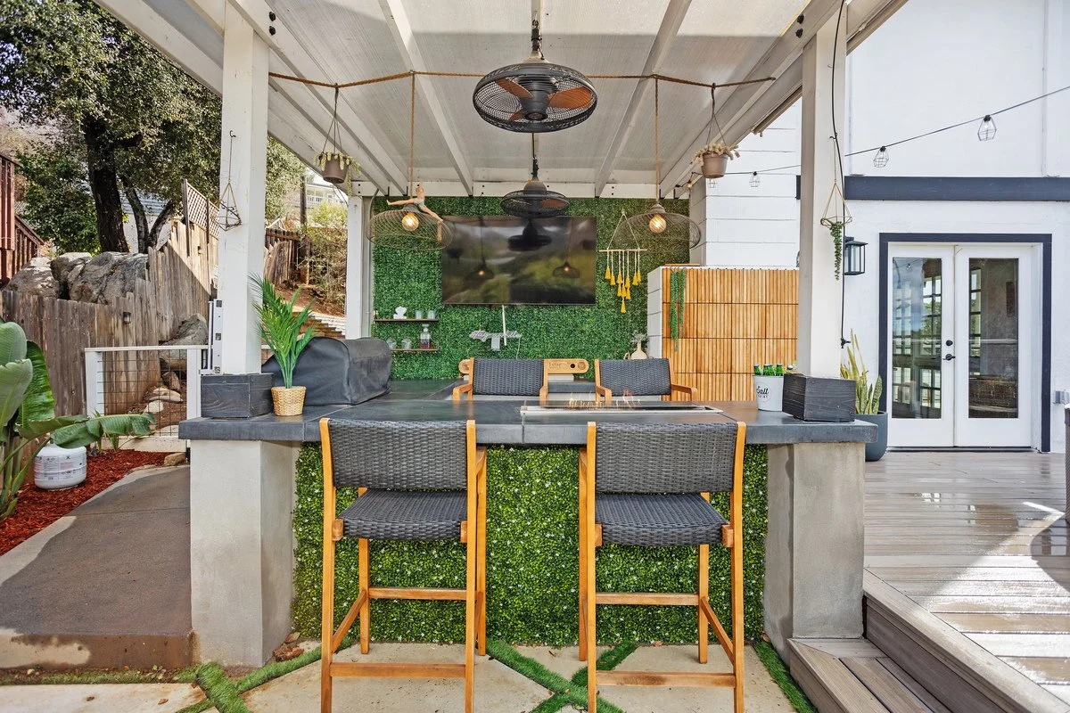 Outdoor patio area with a bar counter, barstools, a TV on a green wall, ceiling fans, hanging lights, potted plants, and a sliding glass door.