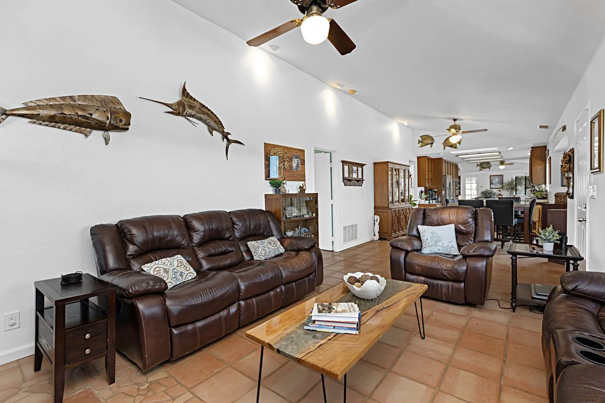 Living room with three brown leather sofas, a wooden coffee table with books and a white bowl, a side table with a remote, wall-mounted fish and marlin decorations, ceiling fans, and a view into the dining and kitchen area with wooden cabinets.