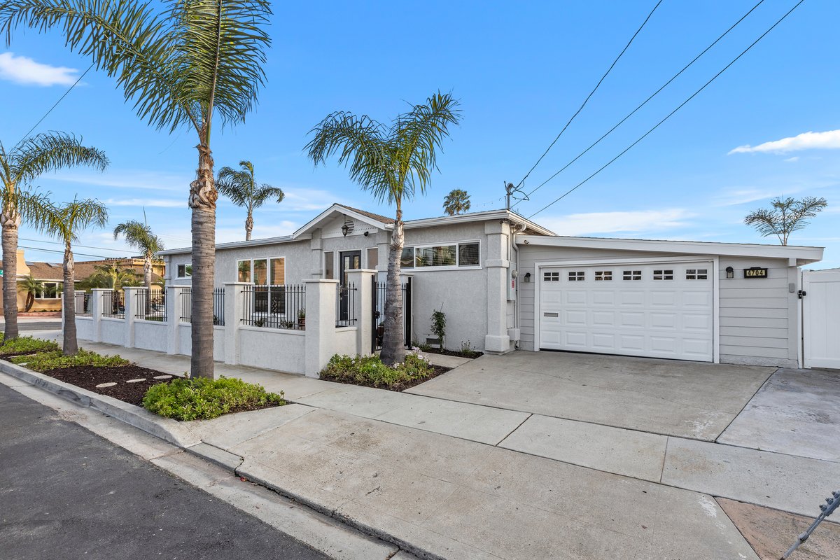 A modern white house with palm trees in the front yard, a white fence, and a two-car garage under a blue sky.
