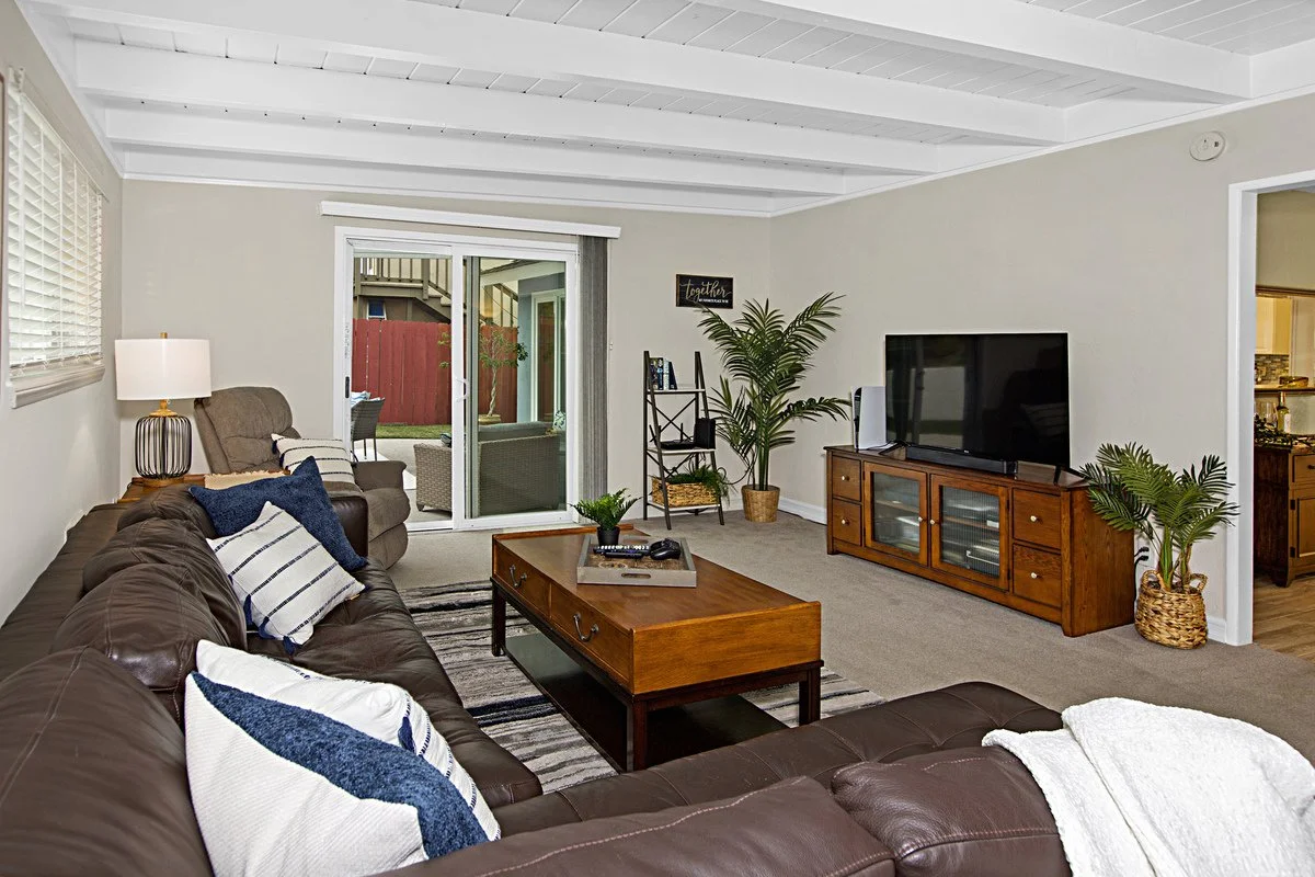 Living room with a brown leather couch, a brown armchair, a wooden coffee table, a TV on a wooden stand, potted plants, a sliding glass door, and window blinds.