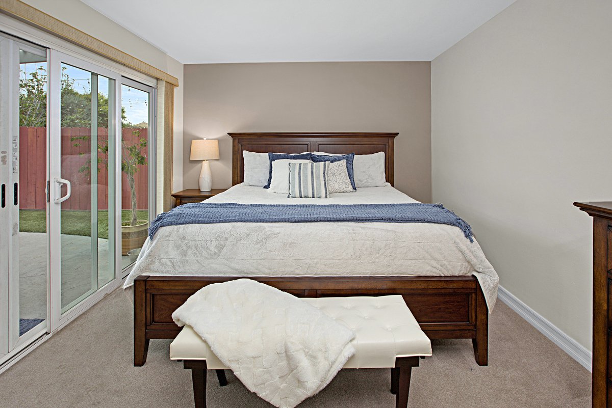 A bedroom with a large bed with a wooden headboard, white bedding, and blue accent pillows. There is a sliding glass door to the outside yard with a tree and a red fence, and a nightstand with a lamp on the left side of the bed.