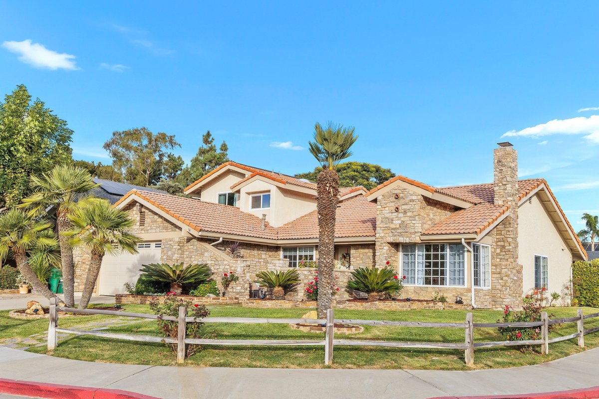 A house with a stone facade, tiled roof, and a chimney, surrounded by palm trees, bushes, and a lawn, under a blue sky with a few clouds.