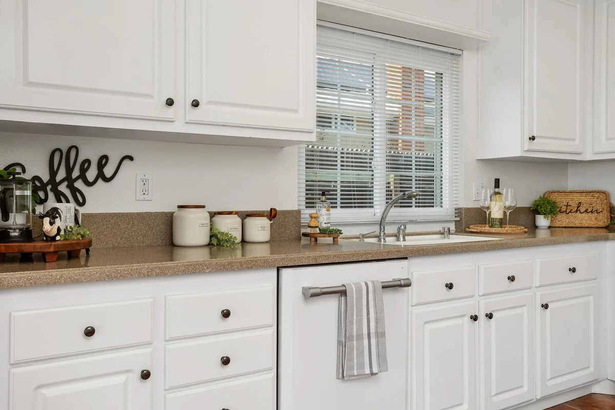 Clean white kitchen with beige countertop, white cabinets, and a window with blinds. Counter has jars, a cutting board, and glasses with wine.