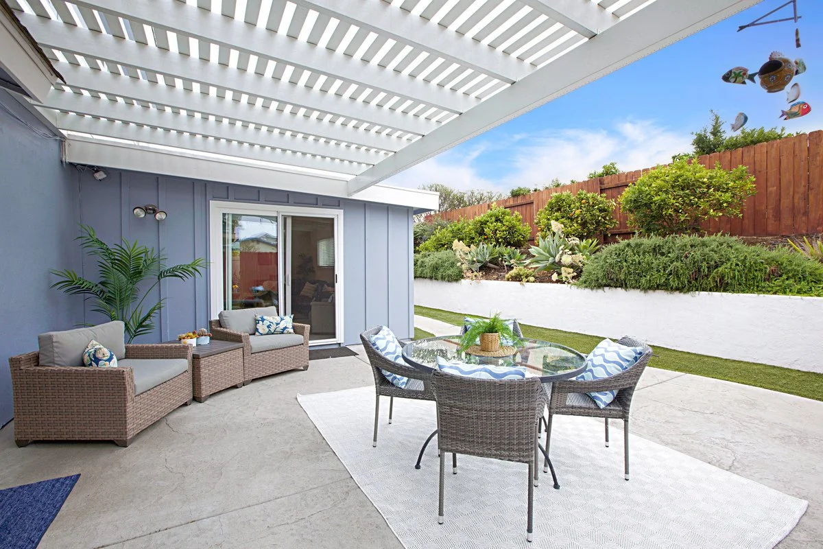 Outdoor patio with wicker furniture, glass table, and porch cover, overlooking a landscaped yard with bushes and trees.