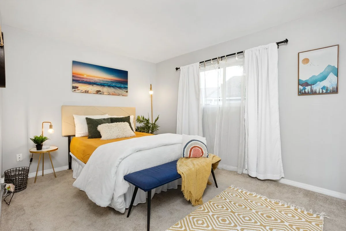 A cozy bedroom featuring a bed with mustard yellow bedding, decorative pillows, and a cream headboard. A small bedside table with a lamp and plant is on the left, while a larger plant is to the right of the bed. A bench with a navy cushion and a yell