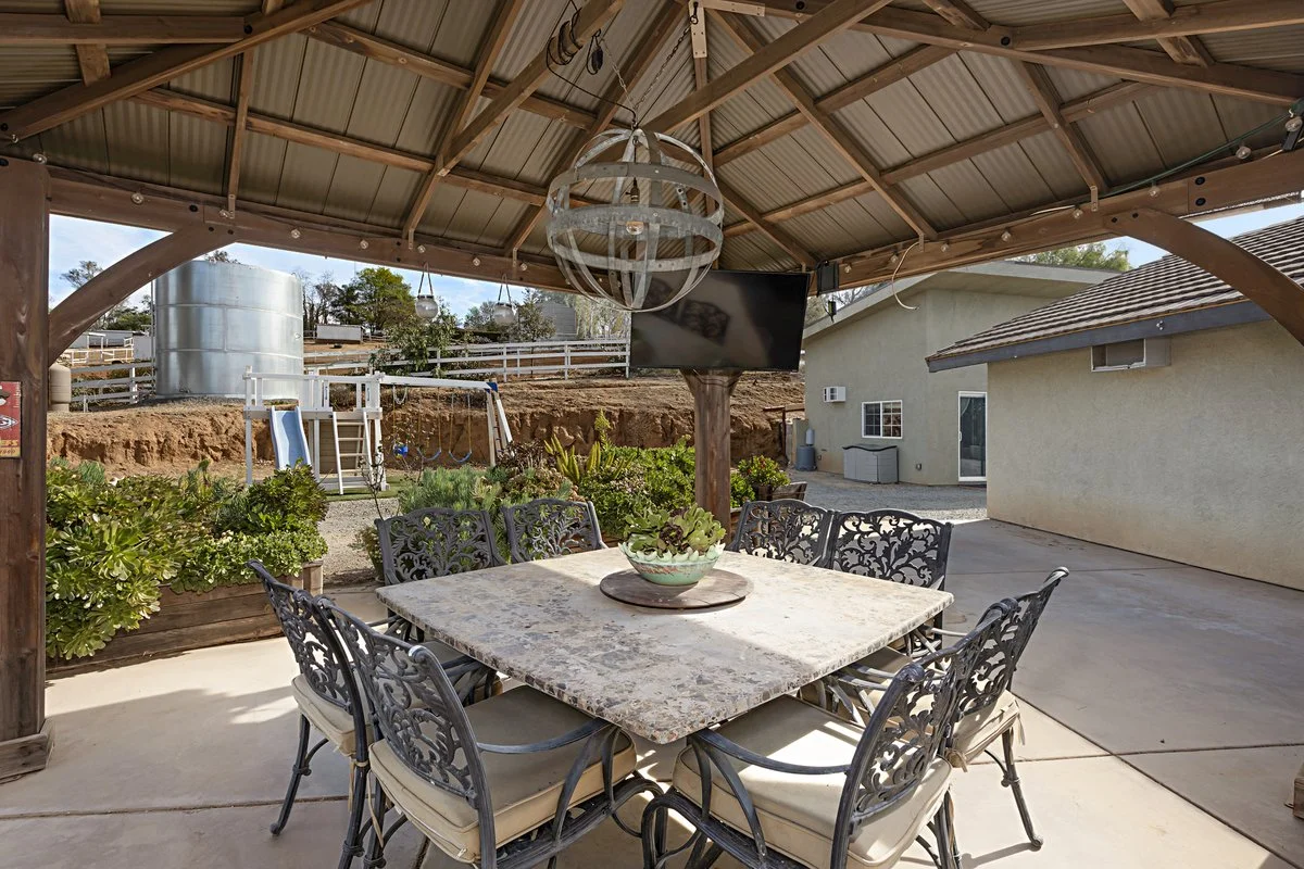 Outdoor patio area with a stone dining table, metal chairs, a hanging spherical chandelier, a wall-mounted TV, and a garden with plants, a slide, and a water tank in the background.