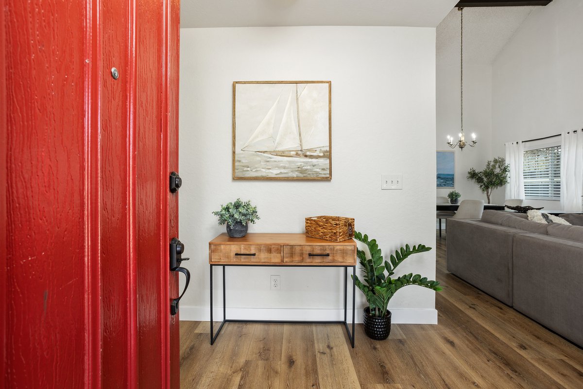 View into a living room from the front door, which is painted red. Inside, there is a wooden console table with two drawers, decorated with a plant and a woven basket. Above the table hangs a framed sailboat painting. To the right, a large L-shaped s