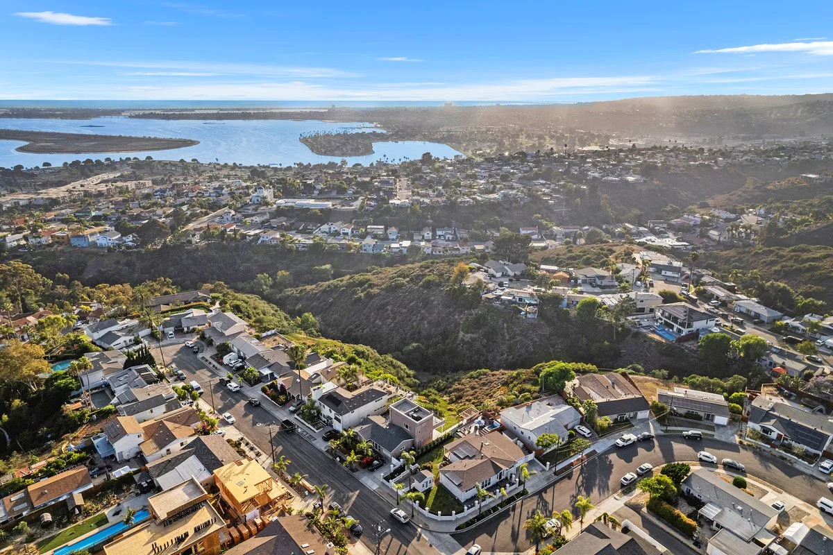 An aerial view of a suburban neighborhood with houses, trees, and streets, overlooking a body of water with islands in the distance under a blue sky.