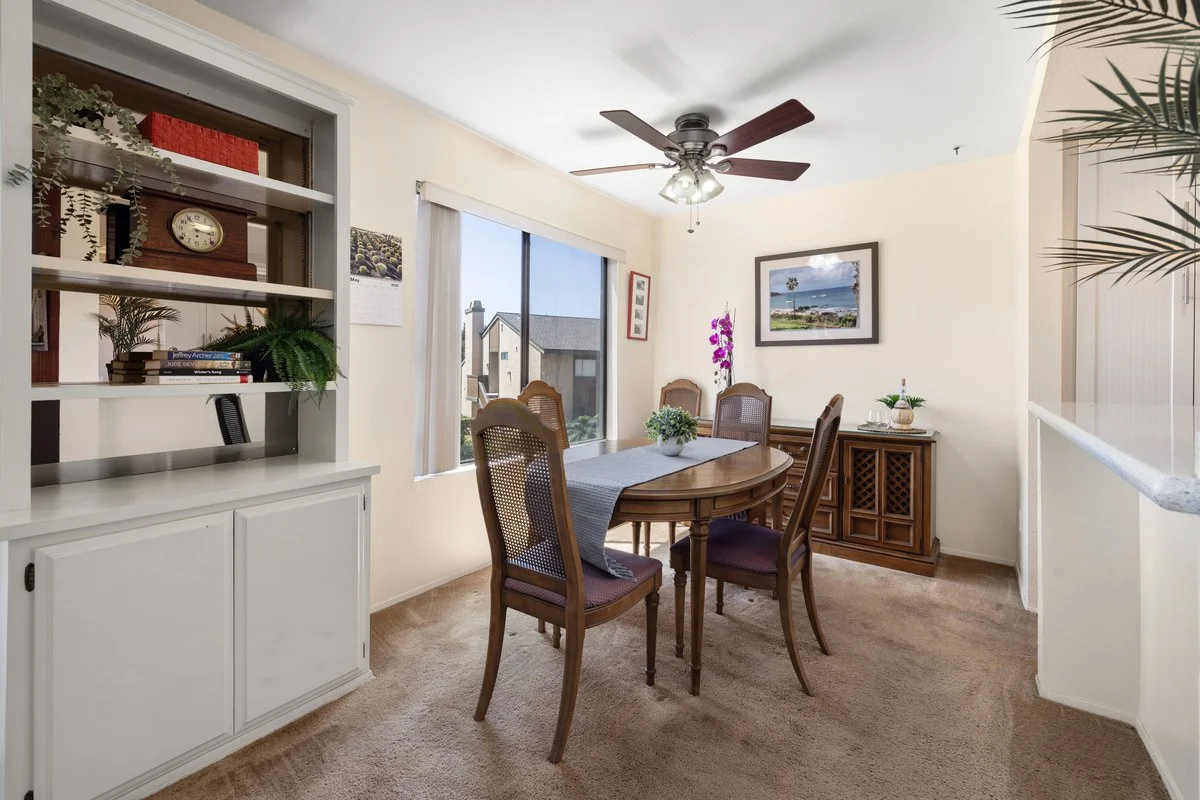 Dining room with a wooden table and six matching chairs, a ceiling fan, a large window with white curtains, a wall hanging picture, and a wooden sideboard with plant decor. A built-in shelf with decorative items and books is partially visible on the 