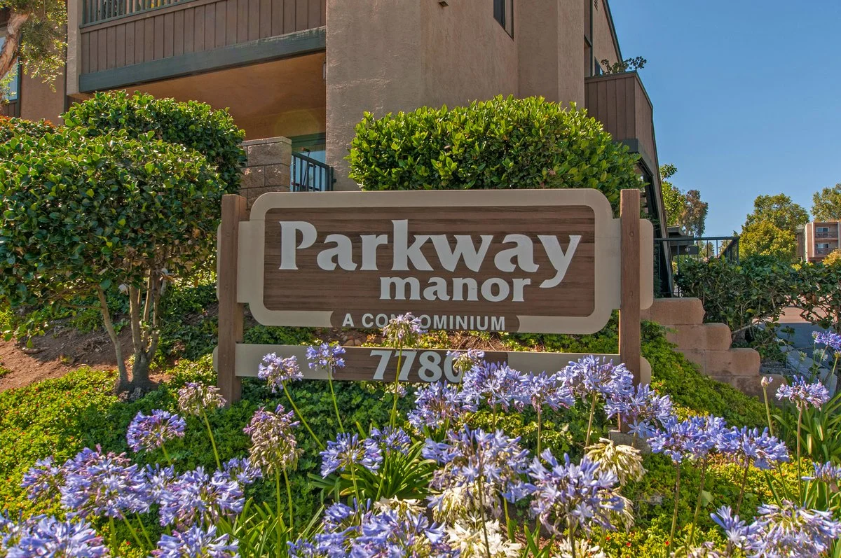 Sign for Parkway Manor apartment complex surrounded by purple flowers and green bushes, with a building in the background.