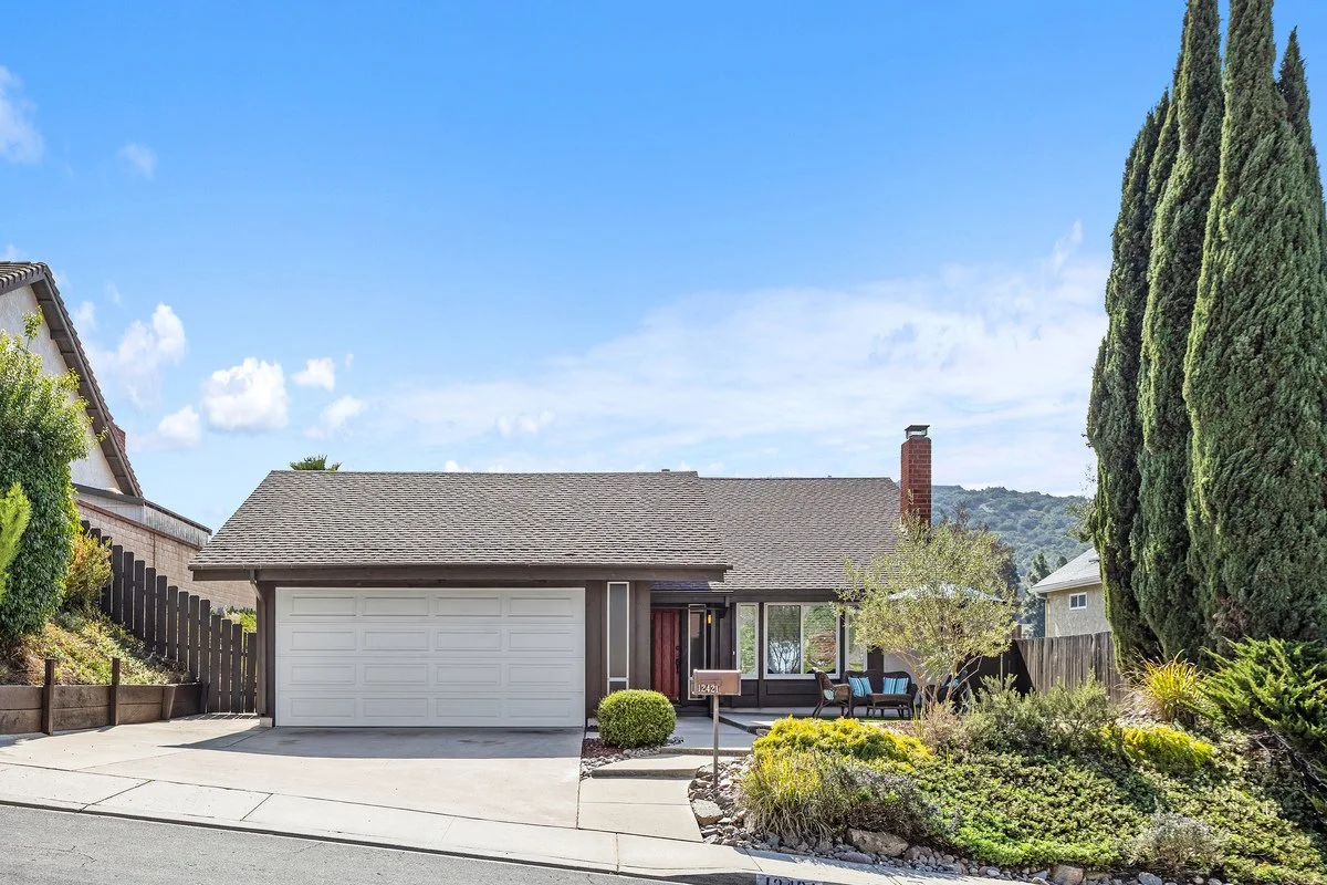 A single-story house with a gray roof, white garage door, and a front porch with outdoor seating, surrounded by landscaping, small trees, and tall cypress trees under a blue sky with a few clouds.
