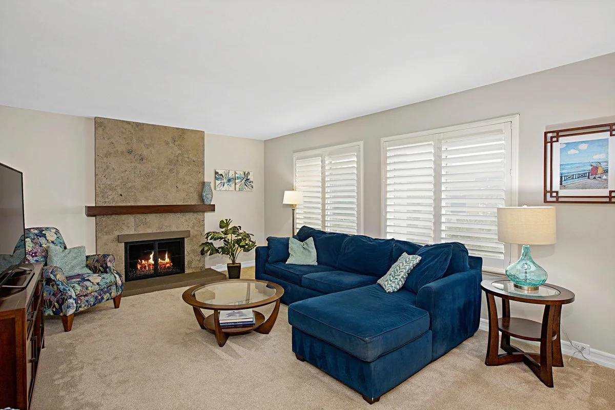 Living room with a blue sectional sofa, a glass coffee table, a floral armchair, a fireplace with a beige stone surround, and three windows with white plantation shutters.