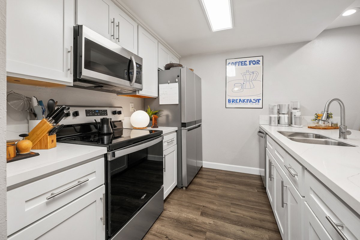 A modern kitchen with white cabinets, stainless steel appliances including a microwave, oven, and refrigerator, and a white countertop with kitchen utensils, a lamp, and a framed coffee-themed poster on the wall.