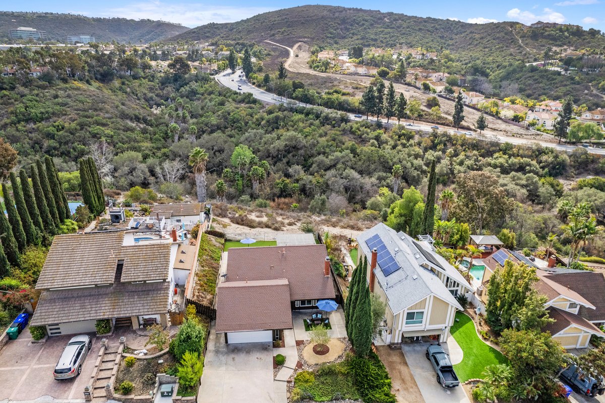 Aerial view of suburban houses with backyards, some with solar panels, amid trees and green landscapes, with a highway on a hillside and distant mountains in the background.