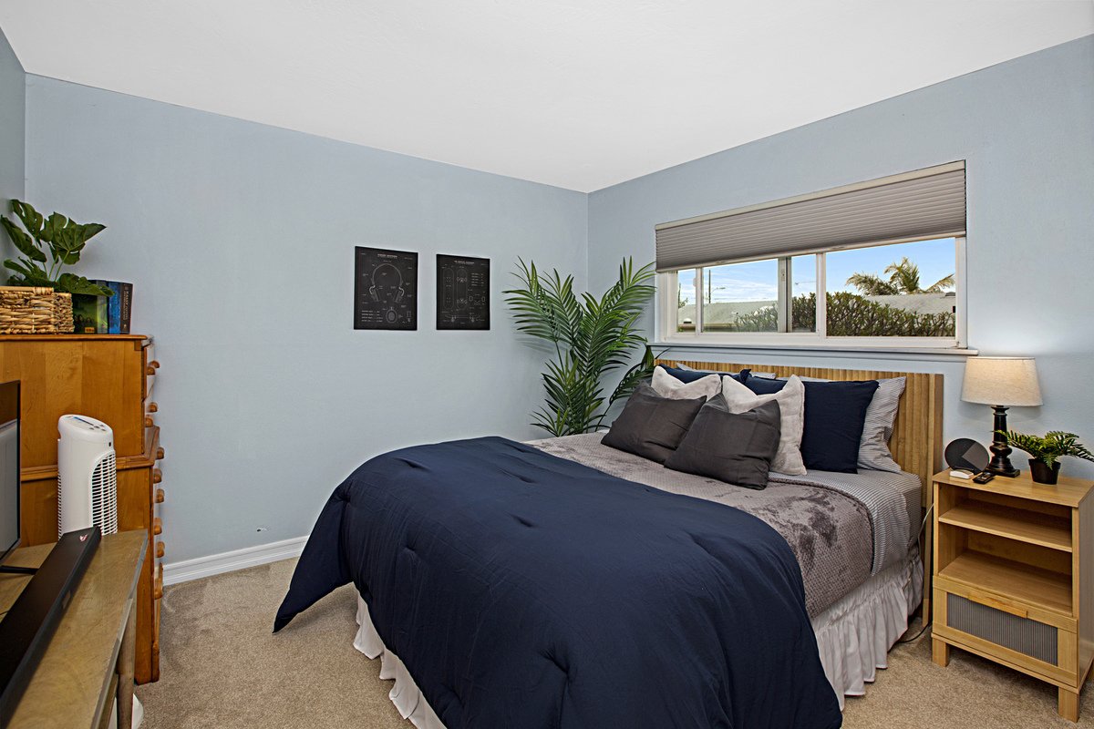 A bedroom with a bed, pillows, a window, a nightstand with a lamp, a plant, a bookshelf, and wall art.