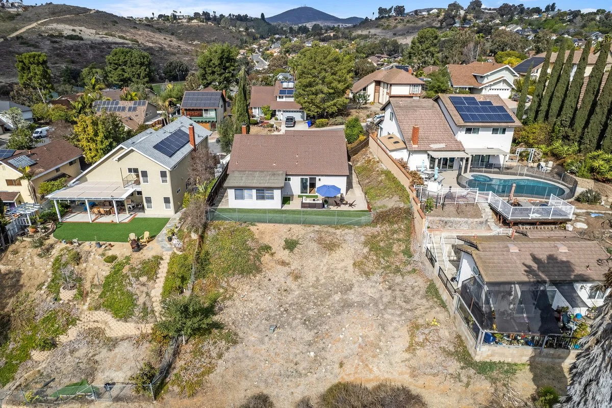 Aerial view of several residential houses with solar panels on the roofs, a backyard with a pool, and a large empty dirt lot in a hilly neighborhood.