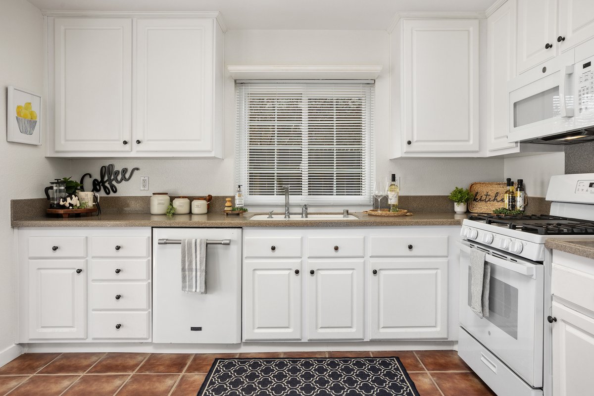 White kitchen with white cabinets, beige countertops, and a window with blinds above the sink. There are various kitchen decor items, including a small plant, a sign that says 'kitchen,' and a bottle of wine. The kitchen has a stove, microwave, and d