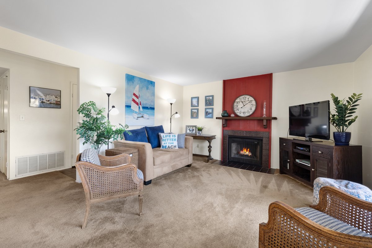 Living room with beige carpet, blue sofa with decorative pillows, paintings of a beach scene, two wicker chairs, a fireplace with a red wall, a large clock, a dark wood TV stand with a flat-screen TV, and potted plants.