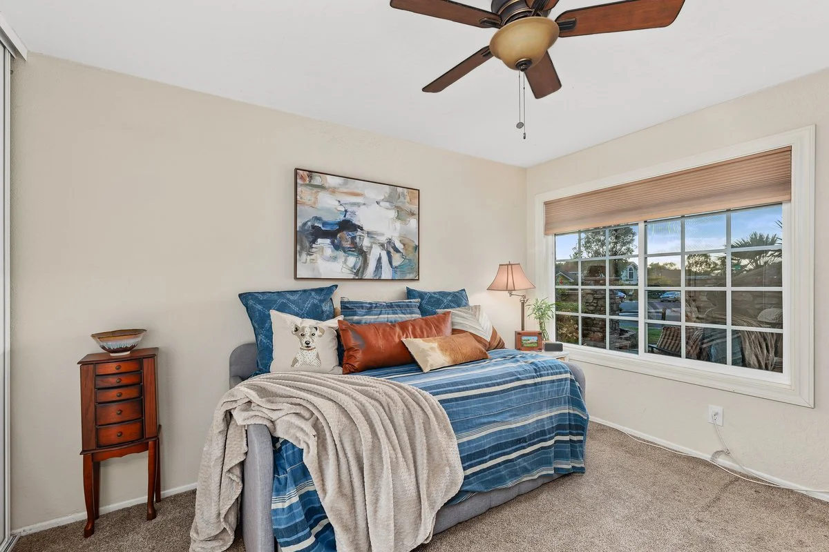 A bedroom with a bed covered in blue striped bedding, decorated with various pillows including a deer pillow, and a beige blanket draped over the side. There is a wooden nightstand with a bowl on it to the left of the bed, and a lamp with a beige sha
