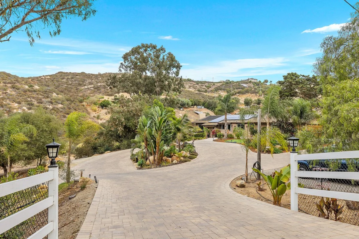 A paved driveway leading to a house, surrounded by desert landscape with trees, shrubs, and mountains in the background, under a blue sky with some clouds.