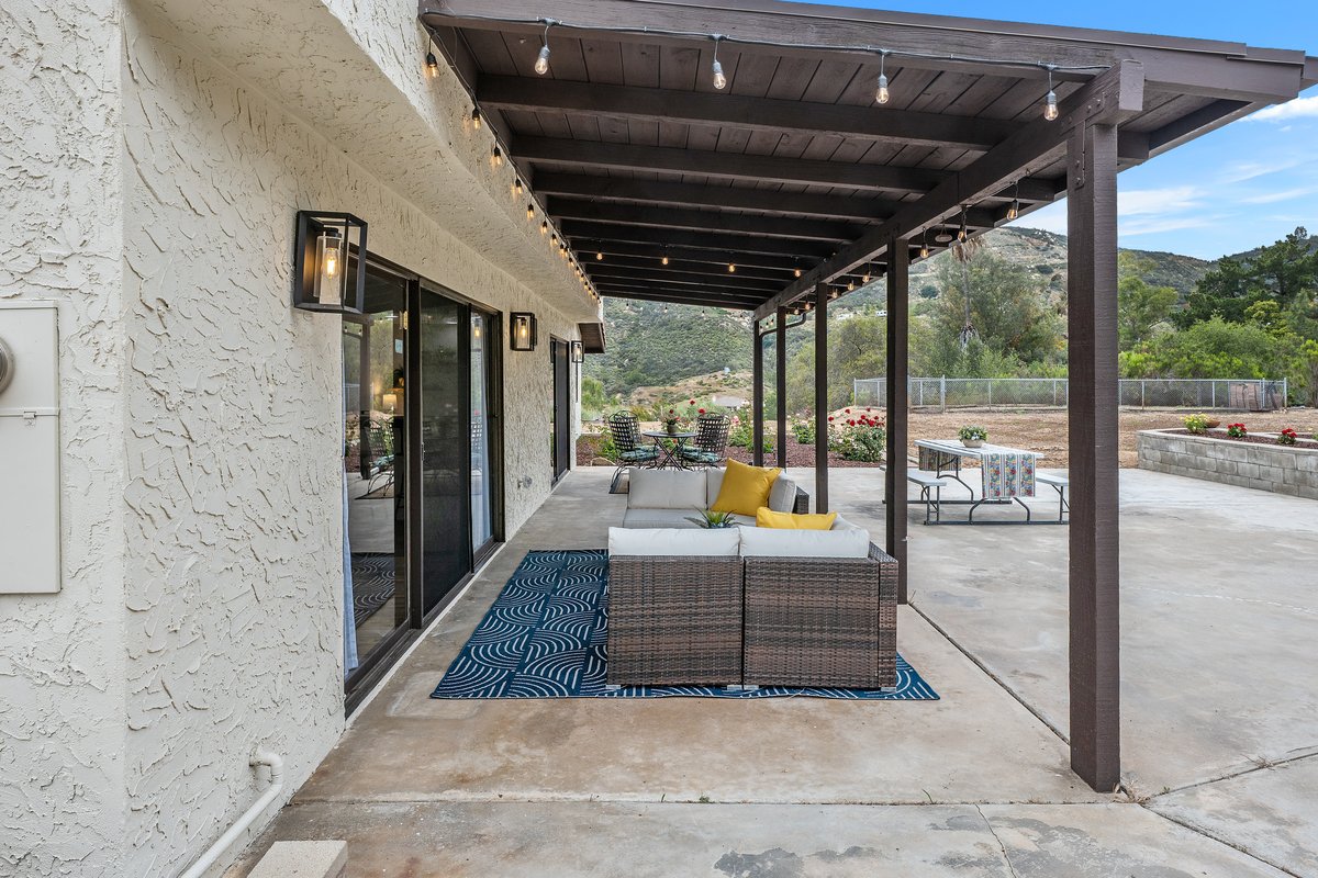 Outdoor patio with a sofa and yellow pillows, string lights hanging from the wooden ceiling, sliding glass doors leading inside, a table with chairs, and a scenic view of mountains and greenery in the background.