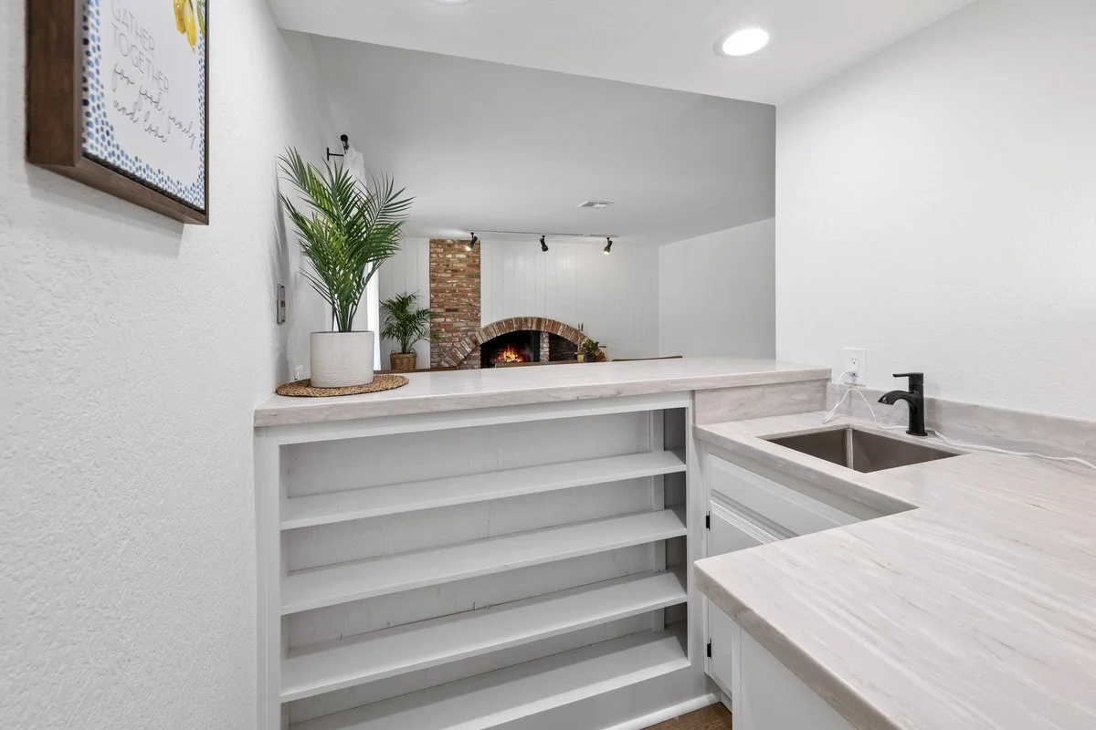 A white kitchen counter with a black faucet and a potted plant, overlooking a living room with a brick fireplace and white paneling on the wall.