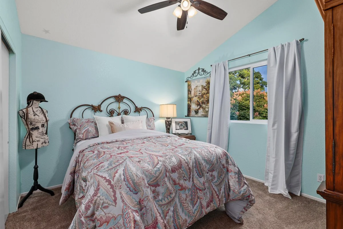A cozy bedroom with light blue walls, a bed with a paisley patterned quilt, decorative pillows, a vintage metal headboard, a nightstand with a lamp, framed photos, and paintings, a window with white curtains showing trees outside, a ceiling fan with 