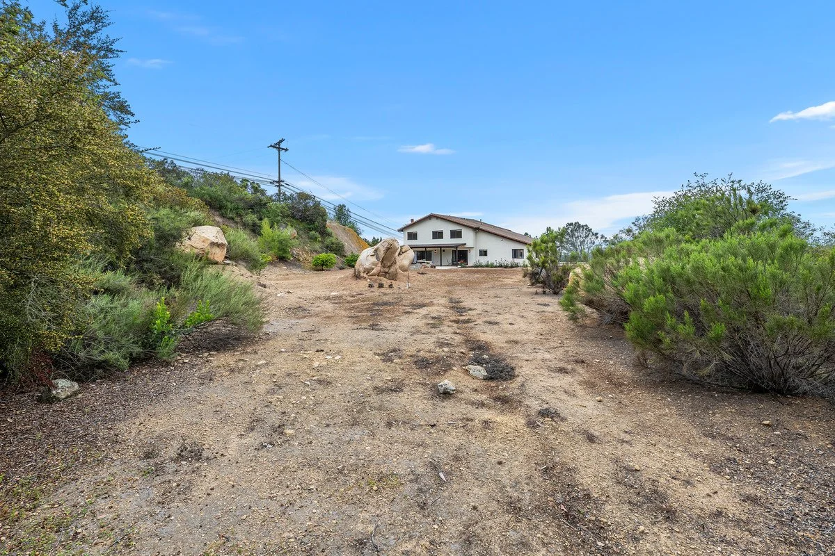 Dirt driveway leading to a house on a hill, with bushes and rocks on either side, under a blue sky with some clouds.