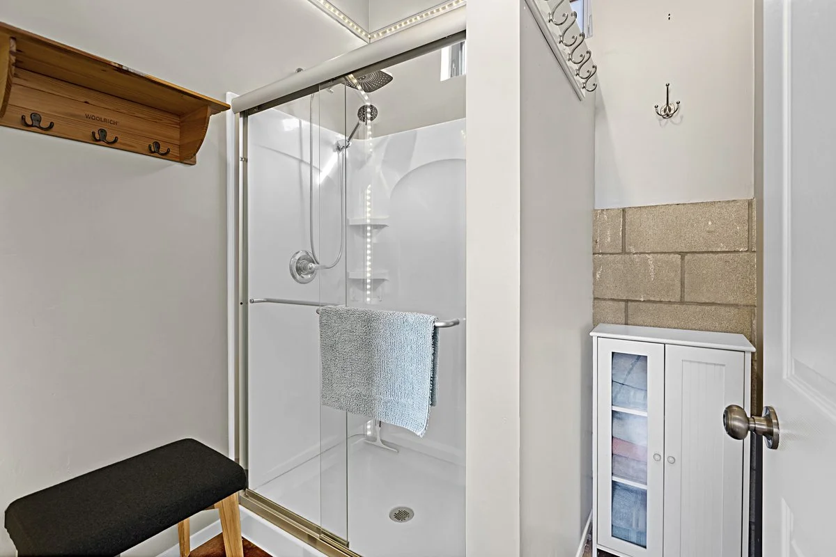 A small bathroom with a walk-in shower with a glass door, a black bench with wooden legs, a wall-mounted wooden coat rack with hooks, and a small white cabinet with glass doors. There is a towel hanging on the shower door handle and hooks on the wall