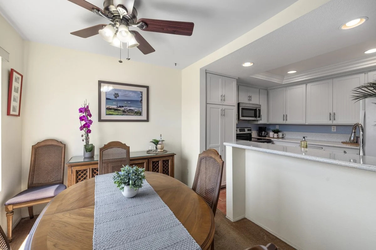 A dining area with a wooden table and four chairs, decorated with a white table runner and a potted plant. A sideboard with decorative items and a framed picture of a beach scene are on the wall. The adjacent kitchen has white cabinets, a microwave, 