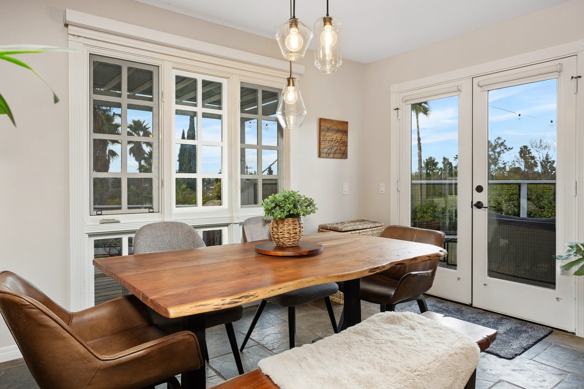A cozy dining area with a wooden table, four chairs, a potted plant centerpiece, and large window and glass door with outdoor view of trees and blue sky.