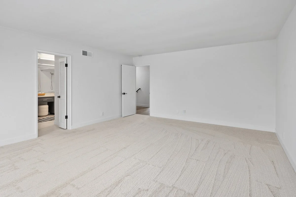 Empty white-walled room with beige carpet, door to a bathroom on the left, door to another room in the center, and a staircase visible through the doorway.
