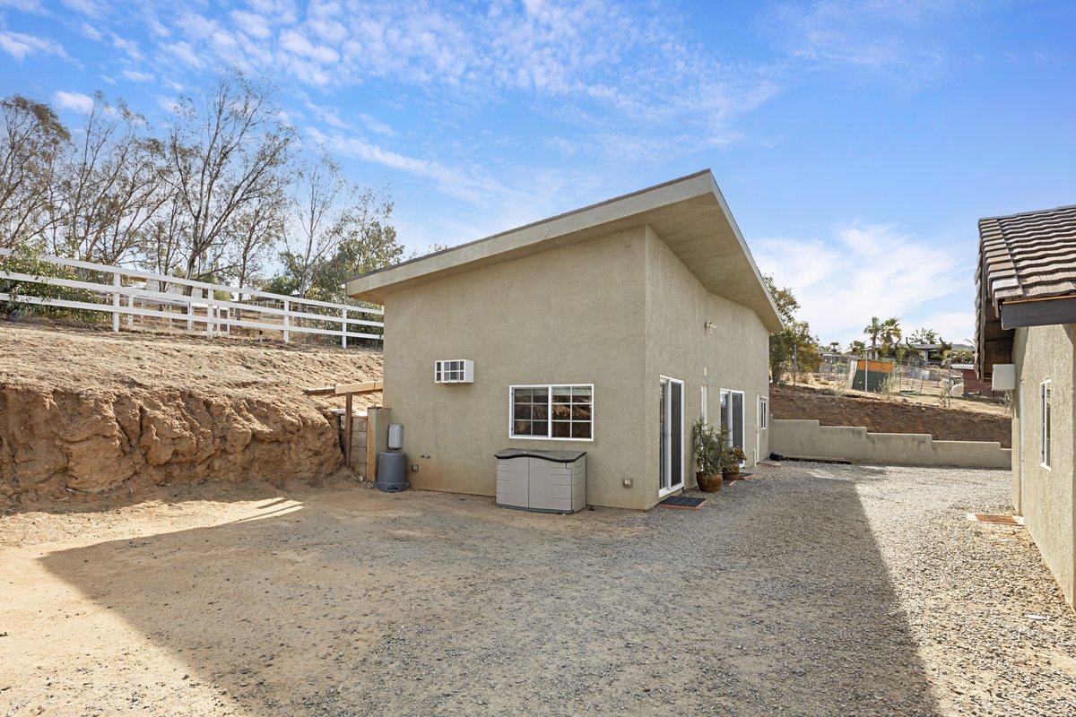 Backyard with beige stucco house, gravel ground, small potted plants, window A/C unit, and a dirt hill with a white fence on top under a blue sky.