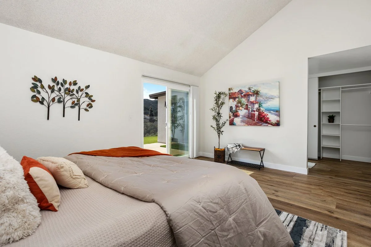 A bedroom with a bed dressed in beige and rust-colored bedding, decorated with pillows, near a sliding glass door leading outside. The room has white walls, wood flooring, and wall art including a metal tree sculpture and a painting of a seaside vill