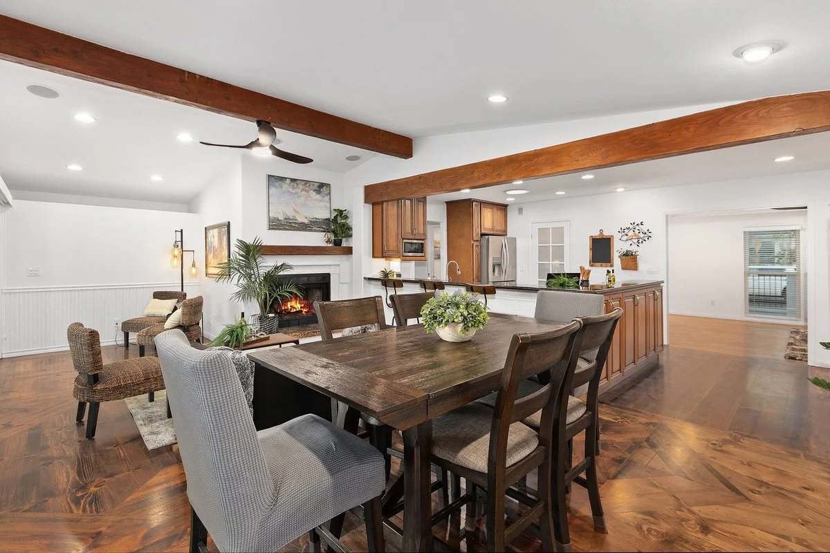 Open-concept living and dining area with hardwood floors, a farmhouse table with chairs, a kitchen with wooden cabinets, stainless steel refrigerator, and a white fireplace, decorated with plants and artwork.