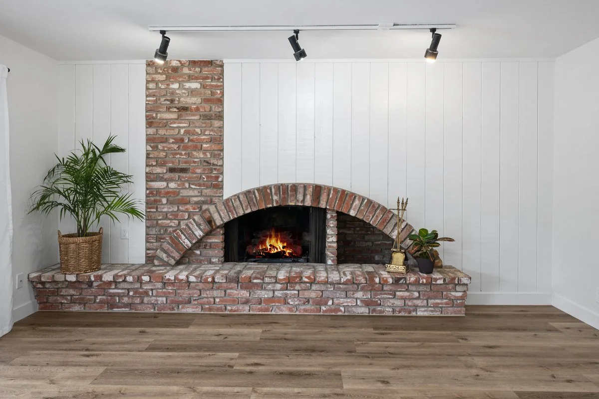 Living room with brick fireplace, white wooden panel walls, potted plant, decorative items, and hardwood floor.