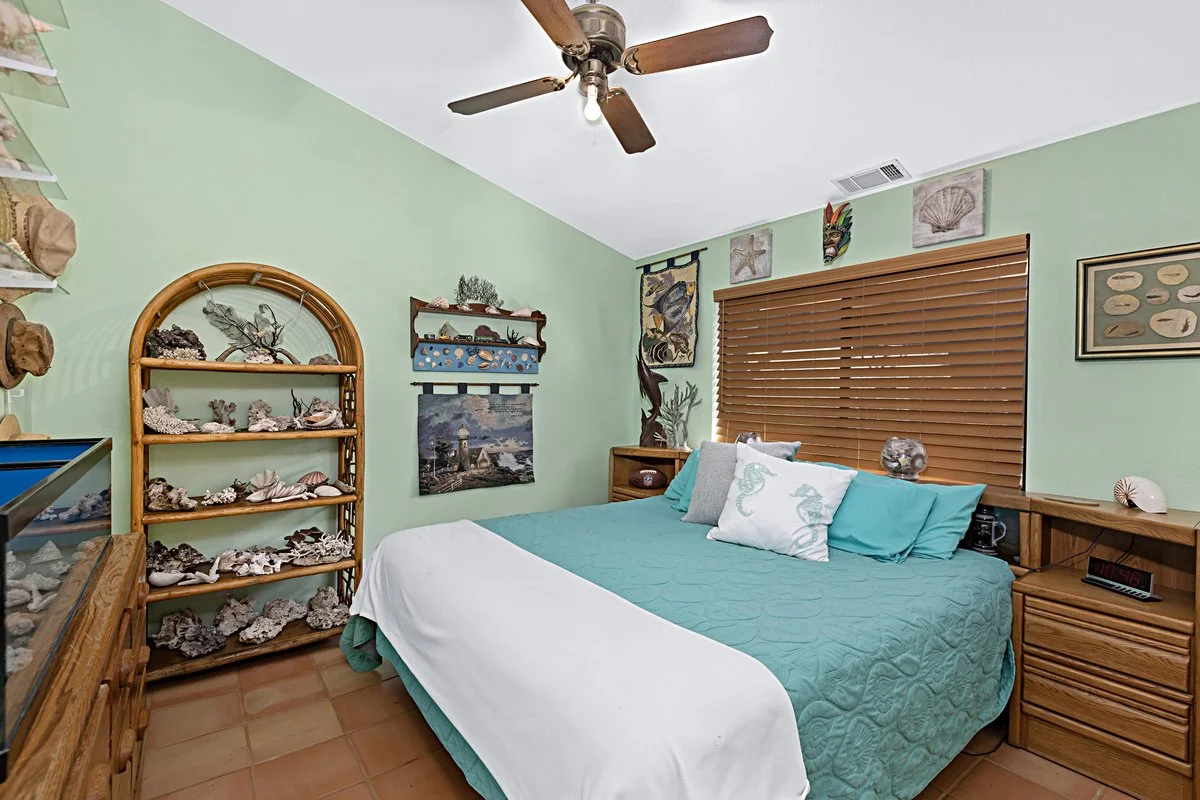 A bedroom with light green walls, a ceiling fan, a window with wooden blinds, a bed with teal bedding, and wooden nightstands. There are decorative items like shells, coral, and ocean-themed pictures on the walls.