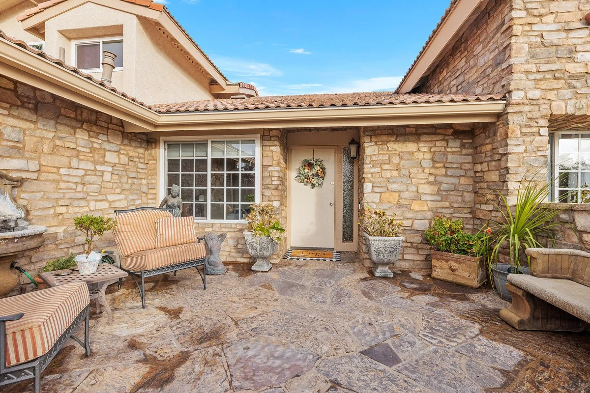 Front porch of a brick house with stone flooring, two striped cushioned benches, potted plants, and a decorative door wreath.