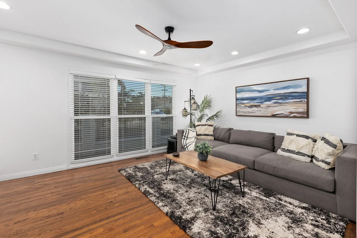 Living room with a large gray sectional sofa, a wooden coffee table, a black and white rug, a painting of a seascape, a ceiling fan, white walls, and a large window with white blinds.