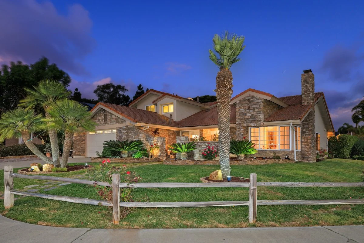 A well-lit house with a stone and stucco exterior, red tile roof, green lawn, palm trees, and a wooden fence in the foreground, during dusk.