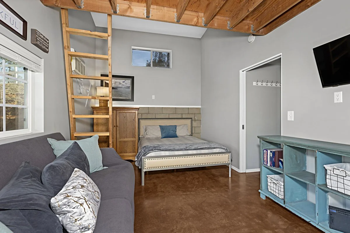 A cozy room with a small bed featuring a beige headboard, a blue pillow, and gray bedding. There is a gray sofa with multiple pillows, a wooden ladder leading to an upper space, a small window above the bed, and a teal shelving unit with baskets and 