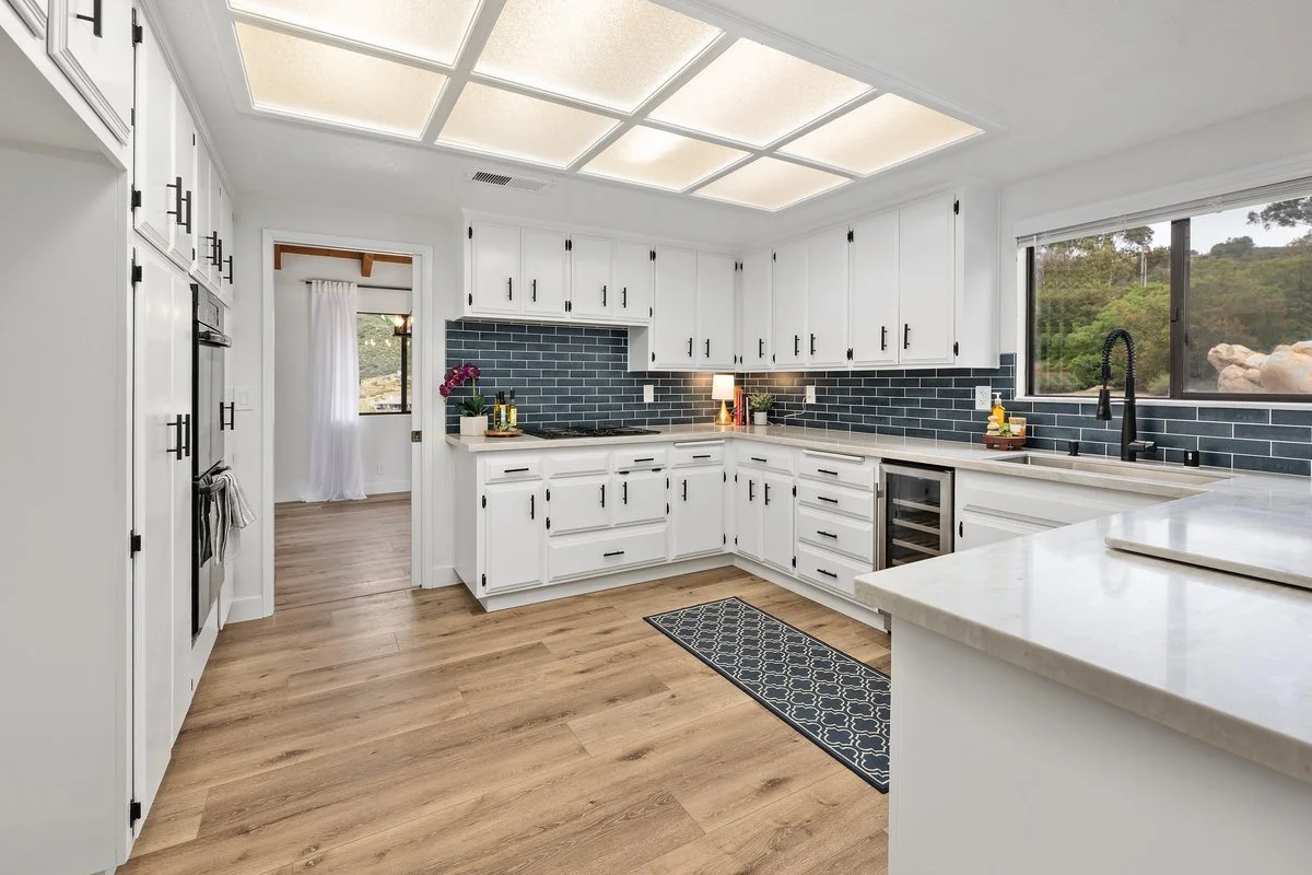 Bright kitchen with white cabinets, black hardware, black tile backsplash, wood flooring, and a large window showing outdoor greenery.
