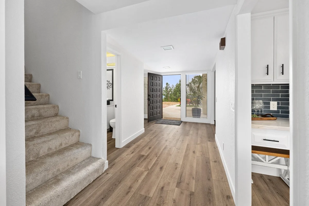 Interior of a modern home entryway with wooden floor, front door open to the outside, staircase on the left, and kitchen visible on the right.